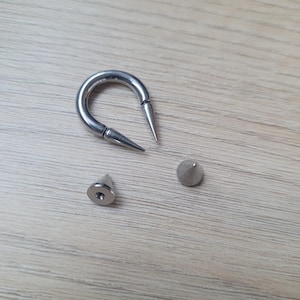 May include: A silver-colored, curved barbell with two pointed ends, along with two separate silver-colored, cone-shaped spikes. The jewelry is displayed on a light brown wooden surface.