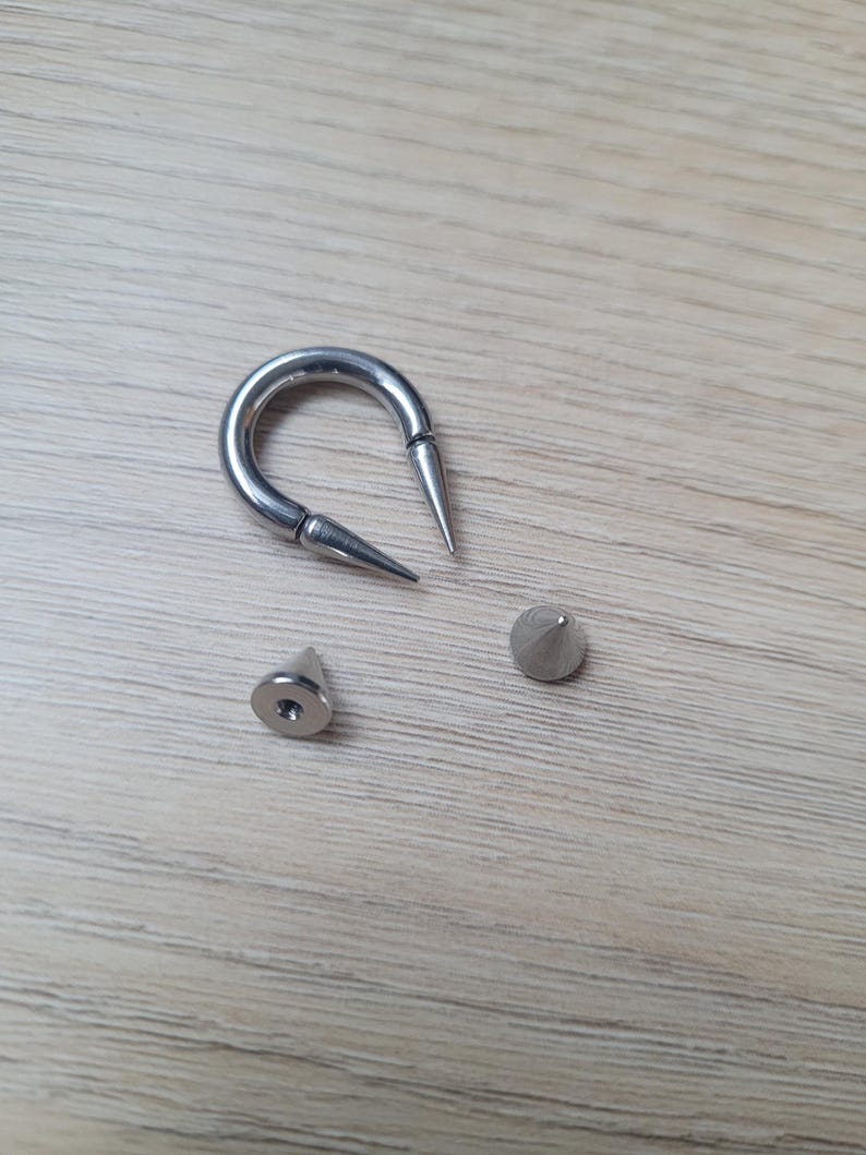 May include: A silver-colored, curved barbell with two pointed ends, along with two separate silver-colored, cone-shaped spikes. The jewelry is displayed on a light brown wooden surface.
