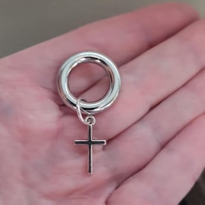 May include: A polished silver-toned circular ear gauge featuring a small, dangling cross charm attached by a thin metal jump ring. The jewelry piece has a smooth, reflective metallic finish and a hollow center, designed for stretched ear piercings.