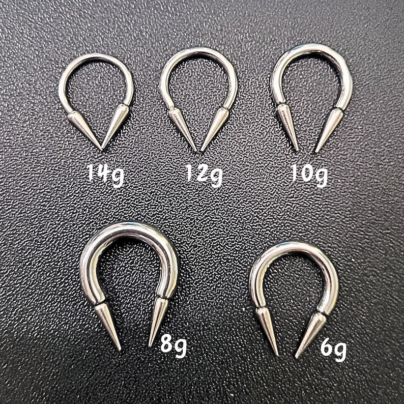 May include: Five silver-colored, U-shaped body jewelry pieces with pointed ends. Sizes are labeled as 14g, 12g, 10g, 8g, and 6g. The jewelry is displayed on a textured, dark surface.