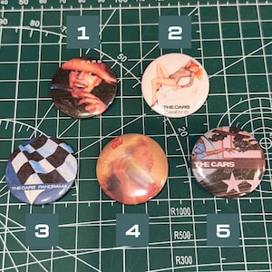 May include: A collection of five round pinback buttons featuring images and text related to the band The Cars. The buttons display various graphics, including album art and band-related imagery. The buttons are on a green cutting mat.