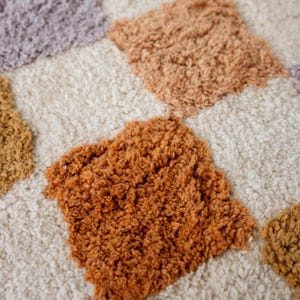 Puede incluir: Primer plano de una alfombra texturizada con un patr&oacute;n de diamantes repetido. Los diamantes son en tonos naranja, beige y morado, sobre un fondo color crema. La alfombra tiene un aspecto suave y afelpado.