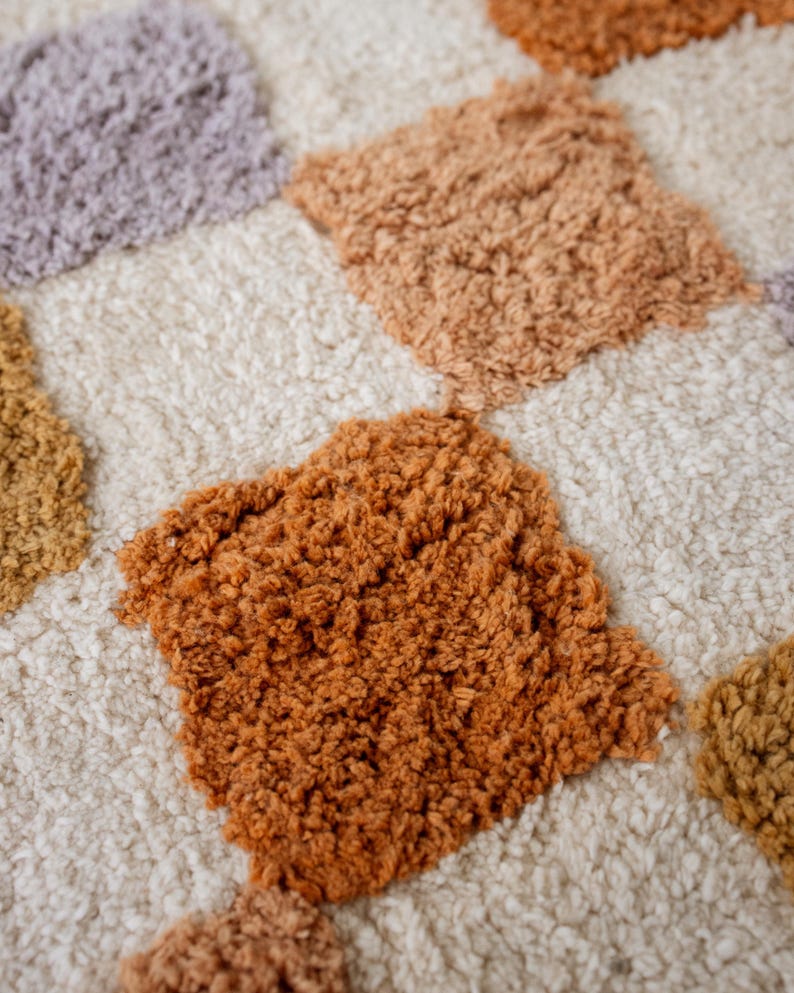 Puede incluir: Primer plano de una alfombra texturizada con un patr&oacute;n de diamantes repetido. Los diamantes son en tonos naranja, beige y morado, sobre un fondo color crema. La alfombra tiene un aspecto suave y afelpado.