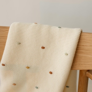 May include: A cream-colored knit blanket featuring a textured garter stitch pattern and small, scattered embroidered dots in muted shades of sage green, terracotta orange, and mustard yellow, draped over a light-toned wooden chair back.