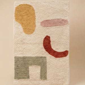 May include: Rectangular rug with a cream-colored background and abstract shapes in mustard yellow, dusty rose, rust, and sage green. The shapes include a large blob, a horizontal oval, a curved line, and a square with a cutout.