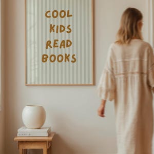 Peut inclure: Affiche encadrée avec le texte "COOL KIDS READ BOOKS" en jaune moutarde. Le fond présente des rayures verticales grises claires et blanches. Un vase blanc est posé sur une petite table en bois avec des livres.