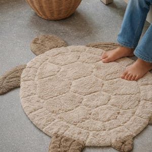 Puede incluir: Una alfombra beige con forma de tortuga con detalles texturizados. La alfombra tiene un cuerpo redondo con cuatro apéndices y un diseño de caparazón estampado. Una cesta de mimbre está en el fondo. La alfombra está diseñada para una habitación infantil.