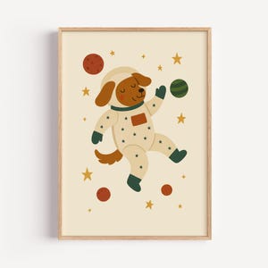 Puede incluir: Una impresión enmarcada que presenta un perro de dibujos animados con un traje espacial, flotando entre estrellas y planetas. El perro es marrón y blanco, con un casco y un traje adornado con estrellas. Los planetas son rojos y verdes, sobre un fondo crema.