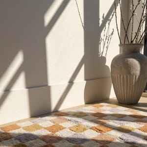 Puede incluir: Una alfombra geom&eacute;trica con dise&ntilde;o de rombos en tonos melocot&oacute;n, naranja, amarillo y lavanda. Un jarr&oacute;n texturizado contiene ramas con brotes. La luz del sol proyecta sombras en la pared. Decoraci&oacute;n interior.