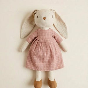 Pode incluir: Boneca de coelho de pelúcia com orelhas compridas e caídas e um vestido rosa xadrez. O coelho tem um nariz castanho claro e sapatos castanhos. A boneca é feita para crianças e tem cerca de 30 cm de altura.