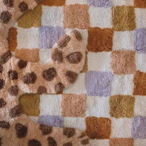 Puede incluir: Una alfombra suave y estampada con un dise&ntilde;o de tablero de ajedrez en tonos blancos, beige, marr&oacute;n y lavanda. Tambi&eacute;n se ve una alfombra separada con forma de animal y manchas marrones, agregando un toque l&uacute;dico.