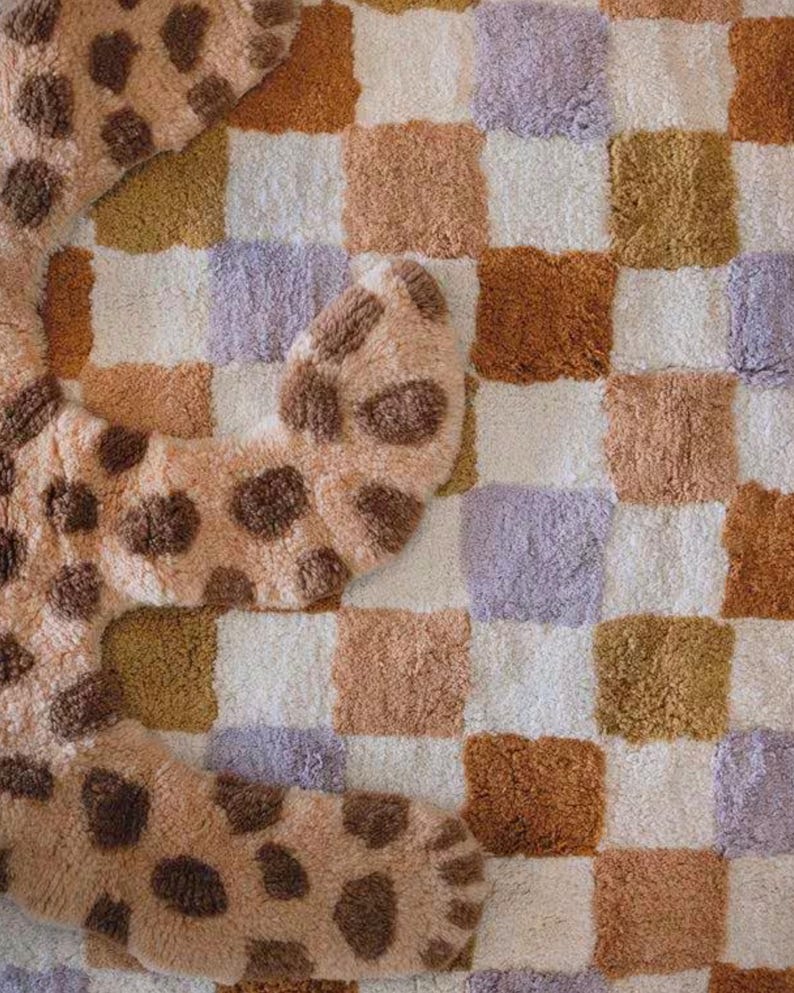 Puede incluir: Una alfombra suave y estampada con un dise&ntilde;o de tablero de ajedrez en tonos blancos, beige, marr&oacute;n y lavanda. Tambi&eacute;n se ve una alfombra separada con forma de animal y manchas marrones, agregando un toque l&uacute;dico.