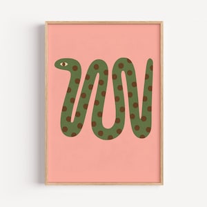Pode incluir: Uma impressão de arte emoldurada com uma cobra verde estilizada com bolinhas marrons em um fundo rosa coral claro. A cobra tem um único olho e um corpo ondulado. A obra de arte está em uma moldura de madeira.