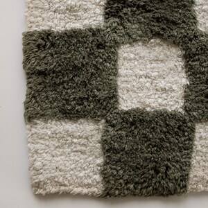 K&ouml;nnte beinhalten: Nahaufnahme eines pl&uuml;schigen, karierten Teppichs. Der Teppich weist ein Muster aus abwechselnden Quadraten in ged&auml;mpftem Olivgr&uuml;n und cremefarbenem Off-White auf. Die Textur wirkt weich und zottelig, was auf einen komfortablen und dekorativen Bodenbelag hindeutet.