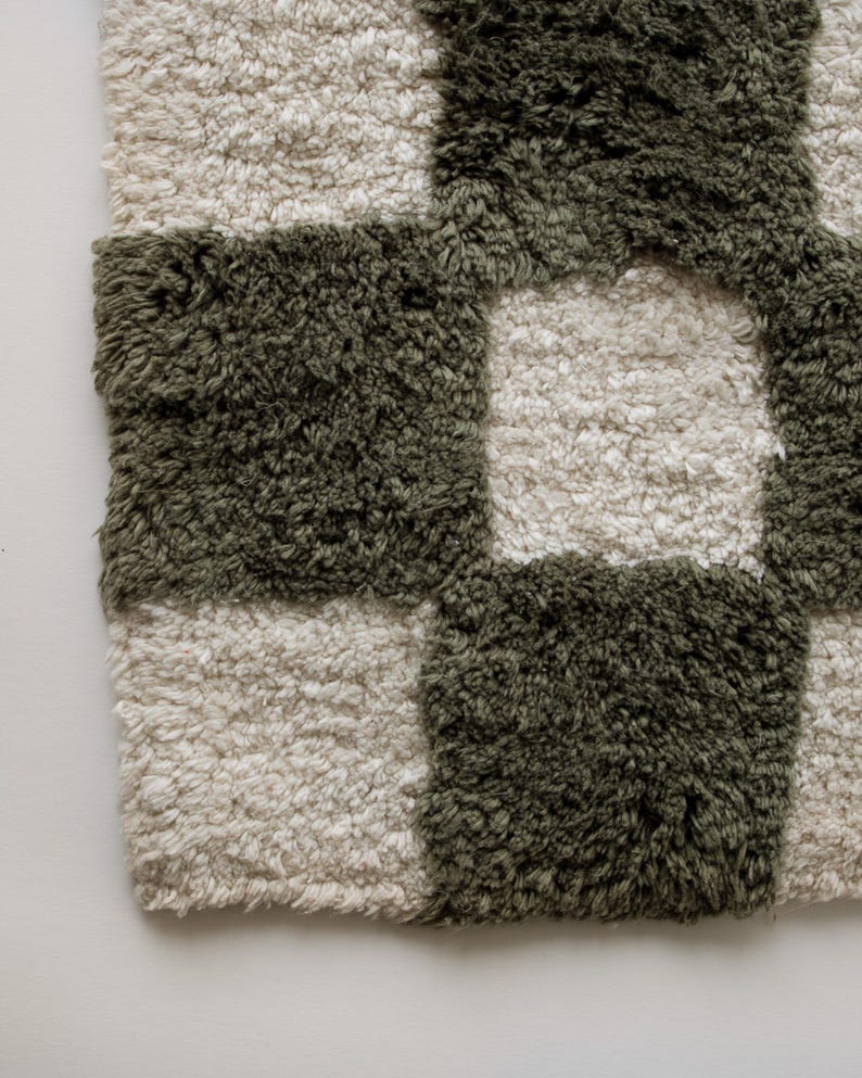 K&ouml;nnte beinhalten: Nahaufnahme eines pl&uuml;schigen, karierten Teppichs. Der Teppich weist ein Muster aus abwechselnden Quadraten in ged&auml;mpftem Olivgr&uuml;n und cremefarbenem Off-White auf. Die Textur wirkt weich und zottelig, was auf einen komfortablen und dekorativen Bodenbelag hindeutet.