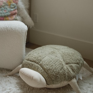 May include: A soft, olive green turtle-shaped cushion with a textured shell pattern. The cushion has four beige legs and a beige head. It is placed on a white, fluffy rug.