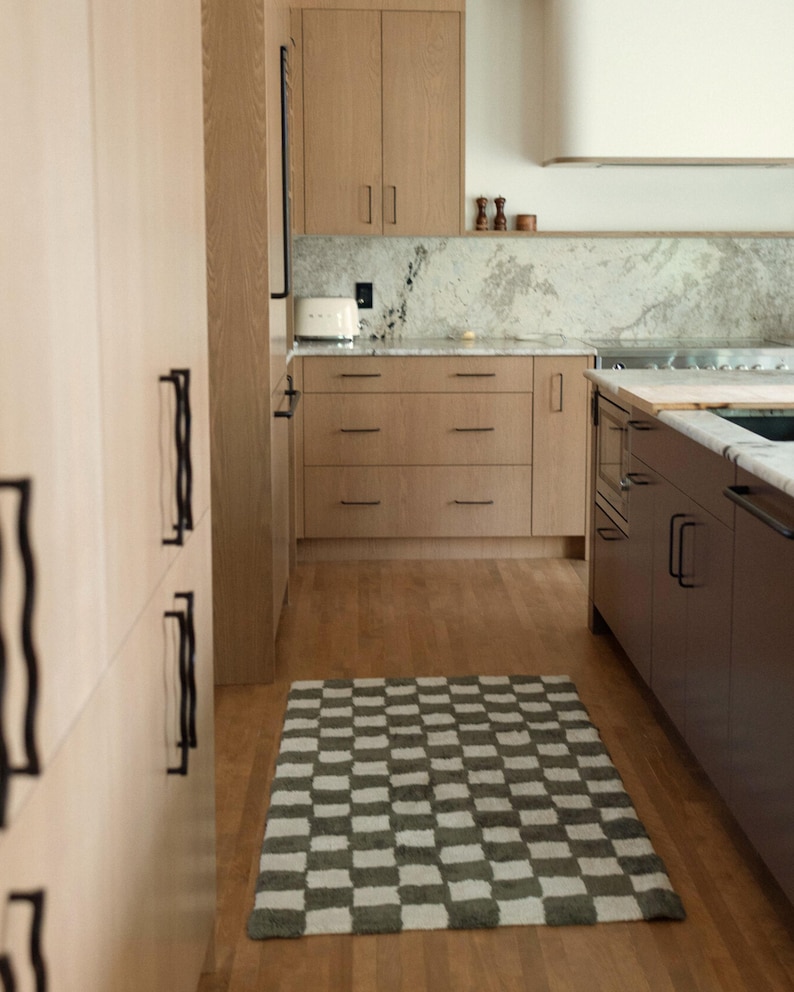 K&ouml;nnte beinhalten: Eine moderne K&uuml;che mit hellen Holzschr&auml;nken und einem karierten Teppich. Die Schr&auml;nke haben schwarze Griffe und die Arbeitsplatte eine Marmorr&uuml;ckwand. Ein wei&szlig;er Toaster steht auf der Theke. Der Teppich ist grau und wei&szlig;.