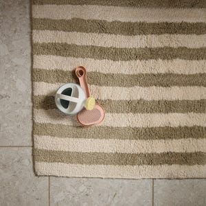May include: A white and green pet grooming ball, a pink brush, and a yellow comb rest on a striped beige and green bath mat. The items are arranged on a light grey tiled floor.