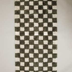 K&ouml;nnte beinhalten: Ein rechteckiger Teppich mit Schachbrettmuster. Der Teppich hat abwechselnd olivgr&uuml;ne und cremefarbene Quadrate. Der Teppich hat ein weiches, strukturiertes Aussehen, was auf ein weiches Material hindeutet. Der Teppich ist f&uuml;r die Inneneinrichtung konzipiert.