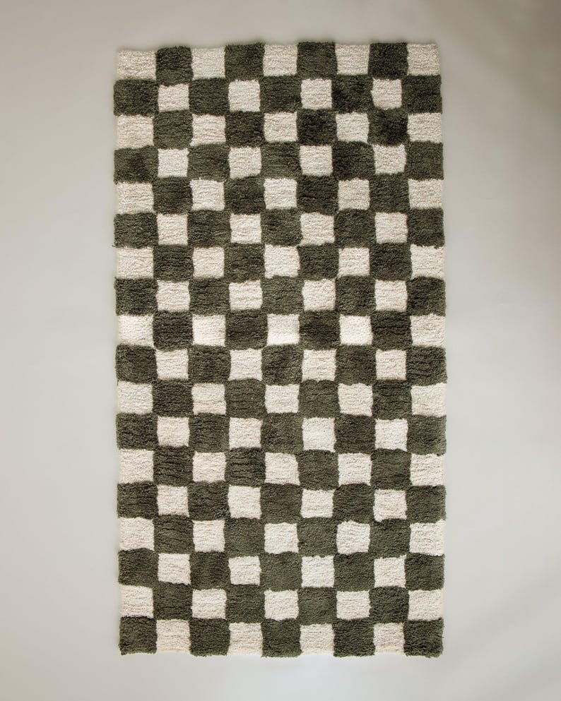 K&ouml;nnte beinhalten: Ein rechteckiger Teppich mit Schachbrettmuster. Der Teppich hat abwechselnd olivgr&uuml;ne und cremefarbene Quadrate. Der Teppich hat ein weiches, strukturiertes Aussehen, was auf ein weiches Material hindeutet. Der Teppich ist f&uuml;r die Inneneinrichtung konzipiert.
