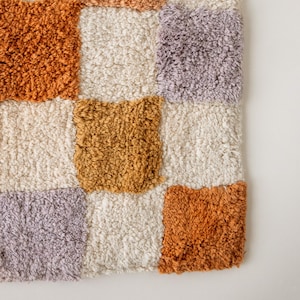 Puede incluir: Primer plano de una alfombra texturizada a cuadros. La alfombra presenta cuadrados de varios colores, incluyendo crema, naranja, dorado y lavanda. El material afelpado crea una apariencia suave y acogedora. El dise&ntilde;o de la alfombra es moderno y geom&eacute;trico.