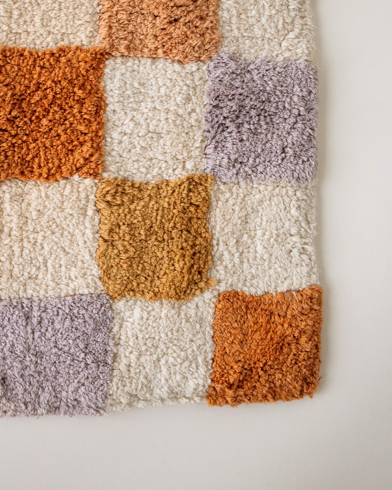 Puede incluir: Primer plano de una alfombra texturizada a cuadros. La alfombra presenta cuadrados de varios colores, incluyendo crema, naranja, dorado y lavanda. El material afelpado crea una apariencia suave y acogedora. El dise&ntilde;o de la alfombra es moderno y geom&eacute;trico.