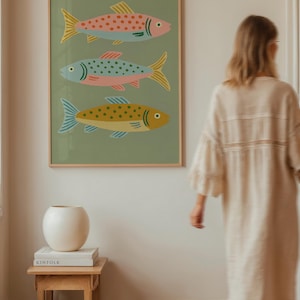 Op de afbeelding: Een ingelijste kunstdruk met drie gestileerde vissen in roze, blauw en geel op een groene achtergrond. De vissen hebben decoratieve patronen. Het kunstwerk bevindt zich in een kamer met een houten bijzettafel en een witte vaas.