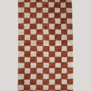 May include: A rectangular rug with a checkerboard pattern. The rug features alternating squares of a warm, reddish-brown color and a neutral off-white. The rug has a soft, textured appearance.
