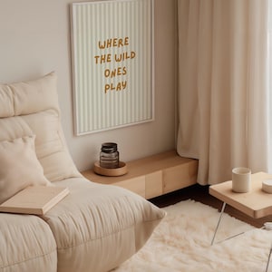 Puede incluir: Un salón acogedor con un sofá crema, una alfombra mullida y una pequeña mesa auxiliar de madera. En la pared, un cuadro enmarcado con el texto "WHERE THE WILD ONES PLAY". También se ven un tarro de cristal y un libro.