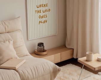 May include: A cozy living room scene with a cream-colored, low-profile sofa, a fluffy rug, and a small wooden side table. A framed print on the wall reads "WHERE THE WILD ONES PLAY." A glass jar and a book are also visible.