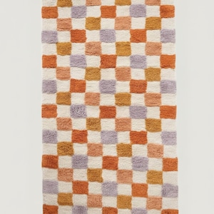 Puede incluir: Una alfombra rectangular con un patr&oacute;n de tablero de ajedrez. Los cuadrados alternan entre blanco roto, naranja, melocot&oacute;n, dorado y lavanda. La alfombra tiene un aspecto suave y texturizado, lo que sugiere un material afelpado.
