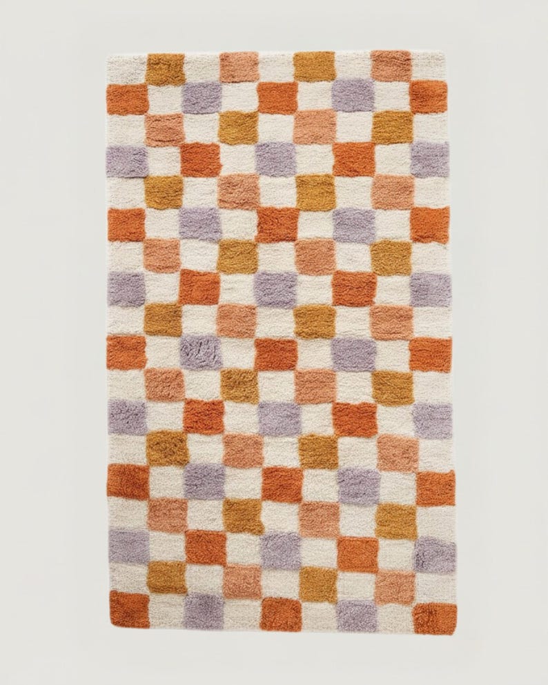 Puede incluir: Una alfombra rectangular con un patr&oacute;n de tablero de ajedrez. Los cuadrados alternan entre blanco roto, naranja, melocot&oacute;n, dorado y lavanda. La alfombra tiene un aspecto suave y texturizado, lo que sugiere un material afelpado.