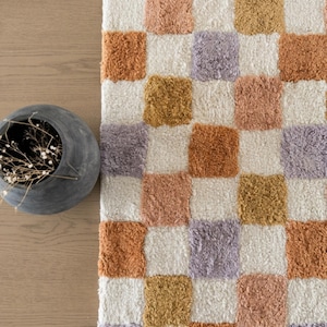 Puede incluir: Una alfombra a cuadros con un patr&oacute;n repetitivo de cuadrados en varios colores, incluyendo blanco, naranja, lavanda y dorado. La alfombra tiene un aspecto suave y texturizado, y est&aacute; colocada sobre una superficie de madera. Un jarr&oacute;n gris con flores secas est&aacute; en la esquina.