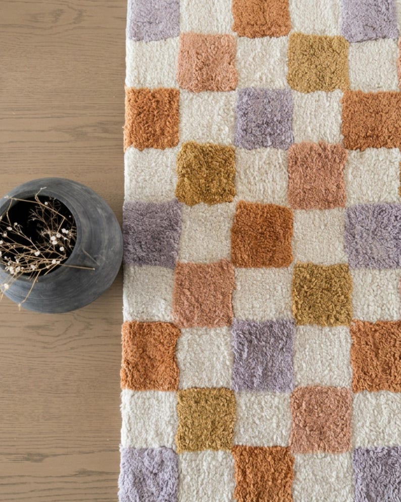 Puede incluir: Una alfombra a cuadros con un patr&oacute;n repetitivo de cuadrados en varios colores, incluyendo blanco, naranja, lavanda y dorado. La alfombra tiene un aspecto suave y texturizado, y est&aacute; colocada sobre una superficie de madera. Un jarr&oacute;n gris con flores secas est&aacute; en la esquina.