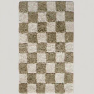 Könnte beinhalten: Ein rechteckiger Teppich mit Schachbrettmuster. Der Teppich hat abwechselnd hellbeige und olivgrüne Quadrate. Der Teppich hat eine weiche, plüschige Textur, was darauf hindeutet, dass er aus einem bequemen Material besteht.