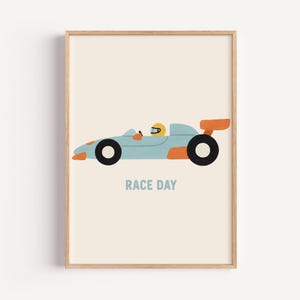 以下が含まれることがあります： 水色とオレンジ色のアクセント、黒い車輪のレーシングカーが描かれたフレームプリント。黄色のヘルメットをかぶったドライバーがコックピットにいます。「RACE DAY」の文字が青色で印刷されています。背景はクリーム色です。