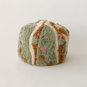 Op de afbeelding: Een ronde, getextureerde poef in tinten groen, bruin, roze en crème. De poef heeft een getuft ontwerp met een herhalend patroon van strepen en stippen. De zachte, pluche textuur suggereert een comfortabele zitoptie.