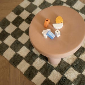 K&ouml;nnte beinhalten: Ein kleiner, runder, pfirsichfarbener Tisch mit einem einzelnen Sockelbein. Auf dem Tisch liegen mehrere bunte Holzbausteine. Der Tisch steht auf einem karierten Teppich mit gr&uuml;nem und cremefarbenem Muster.