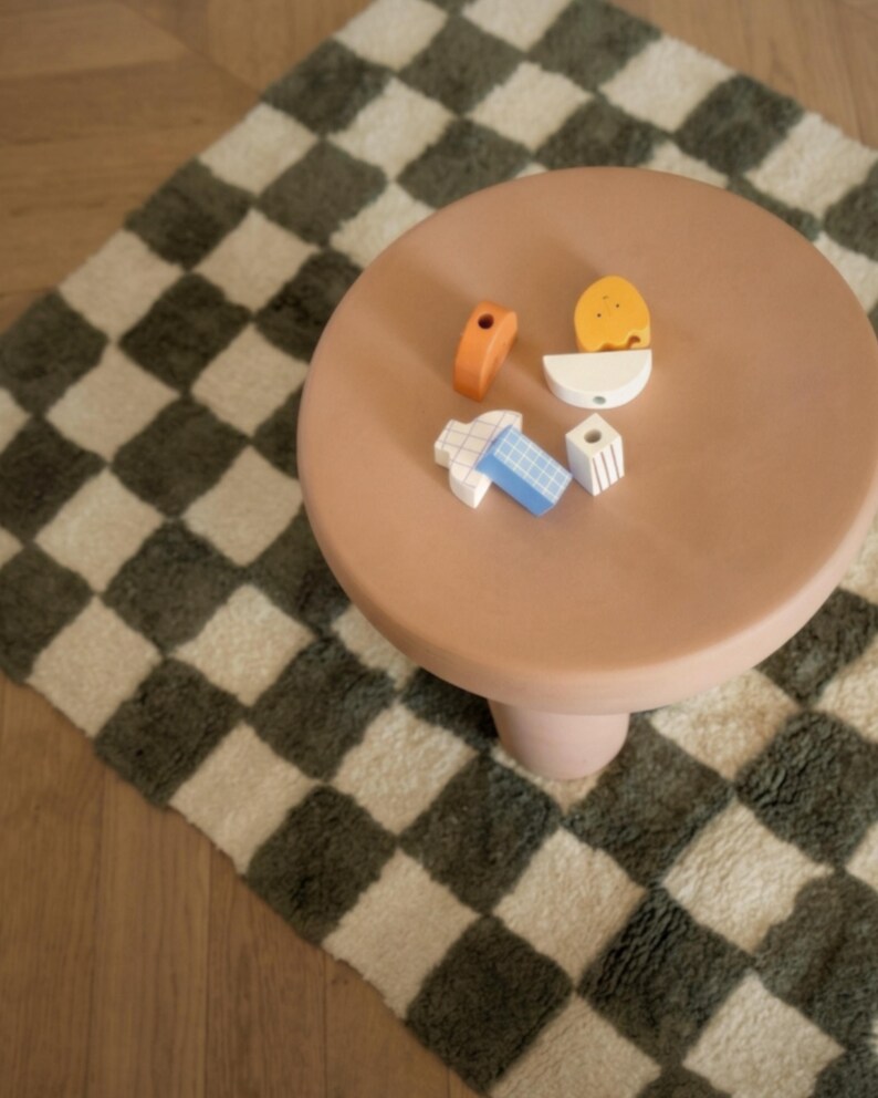 K&ouml;nnte beinhalten: Ein kleiner, runder, pfirsichfarbener Tisch mit einem einzelnen Sockelbein. Auf dem Tisch liegen mehrere bunte Holzbausteine. Der Tisch steht auf einem karierten Teppich mit gr&uuml;nem und cremefarbenem Muster.