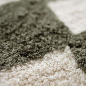 K&ouml;nnte beinhalten: Nahaufnahme eines strukturierten Teppichs mit einem Muster aus olivgr&uuml;nen Formen auf cremefarbenem Hintergrund. Der Teppich hat ein weiches, flauschiges Aussehen mit sichtbaren Schlingenfasern. Das Design ist abstrakt und modern.