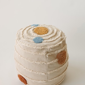 May include: A beige, cylindrical pouf with a textured, fringed design. The top features concentric circles with a mustard-yellow and two blue circular accents. Additional rust-coloured accents are placed on the side, creating a unique, decorative piece.