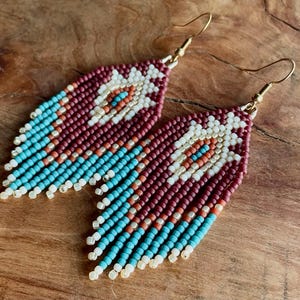 May include: A pair of beaded earrings with a geometric pattern in shades of maroon, white, turquoise, and gold. The earrings have a fringe design and are made with small beads.