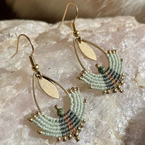 Op de afbeelding: Een paar handgemaakte oorbellen met een macramé-ontwerp. De oorbellen hebben goudkleurige haken en ovaalvormige accenten. Het macramé is in tinten lichtgroen, zwart en bruin, met kleine gouden kralen en een groene steen accent.