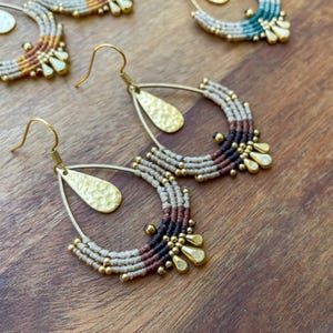 Könnte beinhalten: Handgefertigte Makramee-Ohrringe mit goldfarbenen Tropfenakzenten. Die Ohrringe haben ein halbkreisförmiges Design mit beigen, braunen und schwarzen Makramee-Details. Sie haben goldfarbene Ohrhaken und kleine goldfarbene Perlen.