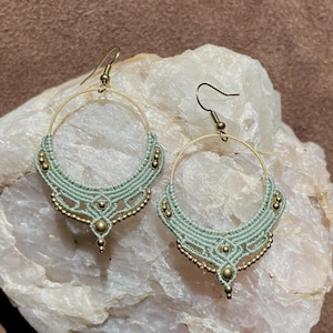 May include: A pair of green and gold hoop earrings with a delicate, intricate design. The earrings are made with macrame and feature small gold beads.