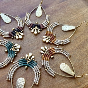 Puede incluir: Colección de pendientes de macramé hechos a mano, de estilo bohemio. Los pendientes presentan un gancho de metal dorado en forma de lágrima y un diseño de semicírculo con cordón tejido en beige, marrón y turquesa. Adornados con cuentas doradas.