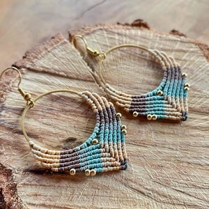 May include: A pair of hoop earrings with a woven design in shades of brown, beige, and turquoise. The earrings have gold-coloured hoops and beads.