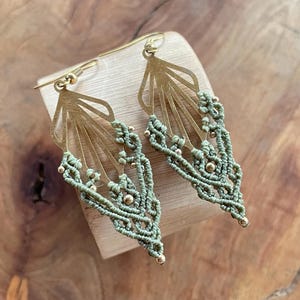 Può includere: Un paio di orecchini fatti a mano con una parte superiore in metallo color oro a forma di ventaglio e intricati dettagli in macramè in un colore verde tenue. Piccole perline dorate sono incorporate nel design in macramè. Gli orecchini sono esposti su un blocco di legno.