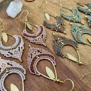 Puede incluir: Primer plano de varios pendientes de macramé en varios colores, incluyendo crema, marrón y verde azulado. Los pendientes tienen forma de lágrima con detalles y ganchos de metal dorado. Se muestran sobre una superficie de madera.