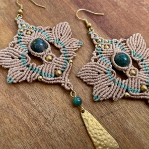 Può includere: Un paio di orecchini in macramè fatti a mano con un accento di pietra verde e un ciondolo dorato. Gli orecchini sono realizzati con cordoncino beige e verde e presentano un design geometrico.